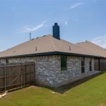 A backyard with a grassy lawn, a wooden fence, and a brick house with a chimney and several windows under a clear blue sky.