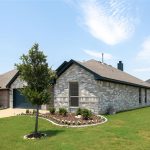 A single-story gray brick house with a dark roof sits on a well-kept green lawn. A small tree and landscaped garden are in the front yard. Neighboring houses are visible on both sides under a blue sky with wispy clouds.