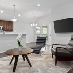 A modern living room with a gray armchair, a chaise lounge, a round coffee table with a white orchid, a mounted TV, a fireplace, and an open kitchen with dark wood cabinets in the background.