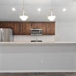 Modern kitchen with dark wood cabinets, stainless steel appliances, a large white island with granite countertop, two pendant lights overhead, and a dining area with a window and chandelier to the right.
