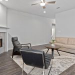 Modern living room with a beige sofa, two black chairs, a round coffee table on a patterned rug, a fireplace with a fire, a wall-mounted TV, and dark wood flooring. Ceiling fan and neutral gray walls.
