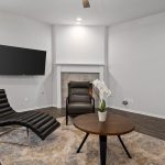 Modern living room with white walls, dark wood flooring, a black lounge chair, a round coffee table with a white orchid plant, a fireplace, a mounted TV, and large windows letting in natural light.