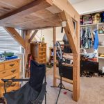 A cluttered bedroom with a wooden loft bed, an open closet full of clothes, shelves with books and bins, a dresser, a folding chair, and various items scattered on the floor and furniture.