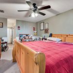 A bedroom with a wooden sleigh bed covered in a red plaid blanket, a ceiling fan with lights, and various framed pictures and decorations on the gray walls. A dresser with personal items is against the left wall.