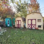 A backyard with a grassy lawn, two yellow sheds with red doors, a small teal building, patio furniture, a grill, trees with autumn leaves, and a wooden fence on the right. Sunlight shines through the trees.
