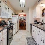 Galley-style kitchen with white cabinets, black countertops, stainless steel appliances, and tile floor. A dining area with a chandelier is visible through a doorway at the far end.
