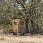 A small, camouflaged hunting blind stands on a wooden platform near the edge of a wooded area, surrounded by trees and dry grass under a clear sky.