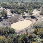 Aerial view of a small, oval-shaped pond surrounded by trees in a rural landscape, with dirt roads and scattered greenery visible around the area.