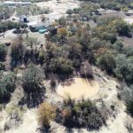Aerial view of a rural landscape with scattered trees, a small muddy pond in the center, and several buildings and water tanks in the background. The area appears dry with patches of sparse vegetation.