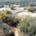 Aerial view of a wooded area with trees in autumn colors surrounding a small, muddy pond and a winding stream, with open fields and more trees in the background.