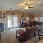 A cozy living room with beige walls, a stone fireplace, brown sofas, a TV, ceiling fan, mounted deer heads, and large windows letting in natural light. A small dog is seen near the glass door on the right.