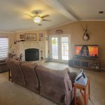 A spacious living room with brown couches, a fireplace, mounted deer heads, a ceiling fan, and a TV showing a woman’s face. Multiple windows with blinds let in natural light; the walls are decorated with pictures and collectibles.