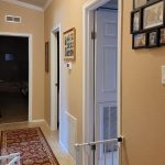 A hallway with beige walls, a red patterned rug, several framed photos, and three doors—one open showing a glimpse of a bedroom. A white baby gate is attached to the wall in the foreground.