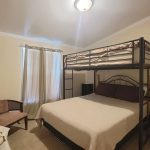A bedroom with a large bed beneath a metal loft bed, a chair, a table with a lamp, curtains covering two windows, and a wall-mounted TV. The room has light-colored walls and carpeted floor.