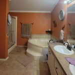 A bathroom with orange walls features a white toilet, a shower, a large corner bathtub, and a double-sink vanity with a wide mirror. Towels hang on the walls, and various toiletries are on the counter.