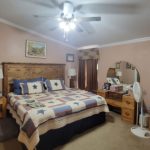 A cozy bedroom with a large bed covered by a star-patterned quilt, wooden headboard, dressers, lamps, a ceiling fan with lights, framed art on pink walls, and brown curtains on the windows.