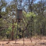 A rusty metal deer feeder stands on tall legs in a dry, rocky area, surrounded by a wire fence and trees with sparse green foliage under a clear blue sky.