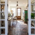 A sunlit room with brick flooring, wooden chairs around a table, potted plants, and a green cabinet. Double glass doors open into the space, and a wooden door with windows is at the far end.
