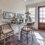A bright sunroom with white paneled walls, brick floor, a rustic table with potted plants, two wooden chairs, a wooden door with glass, large windows, and a distressed turquoise cabinet. Sunlight fills the cozy space.