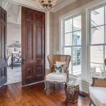 A bright sitting area with two wicker chairs, patterned cushions, a small side table, and large windows letting in natural light, adjacent to a bedroom with dark wooden double doors.