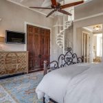 A bright bedroom with a wrought iron bed, a patterned rug, a wooden dresser, a wall-mounted TV, and a spiral staircase leading up. The room features large windows, houseplants, and neutral-toned walls.