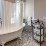 Clawfoot bathtub with white shower curtain, surrounded by marble tile walls and floor. Black metal rack holds folded towels. Large window and white towels hang on wall; bathroom vanity visible in adjoining room.