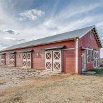 A red barn with white trim and multiple sliding doors stands on a dirt and gravel lot, surrounded by open fields under a partly cloudy sky.