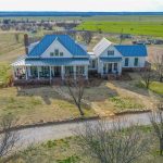 Aerial view of a large farmhouse with a blue metal roof, wraparound porch, and white exterior, surrounded by open fields and scattered trees under a clear sky. Gravel driveway leads to the house.