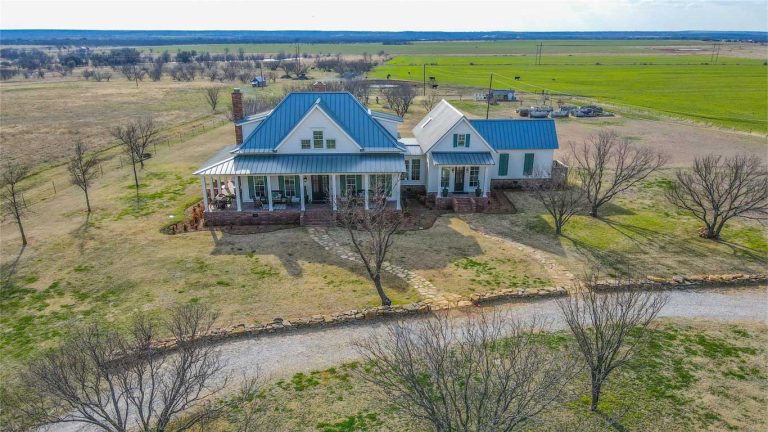 Aerial view of a large farmhouse with a blue metal roof, wraparound porch, and white exterior, surrounded by open fields and scattered trees under a clear sky. Gravel driveway leads to the house.