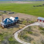 Aerial view of a farmhouse with a large porch and chimney, surrounded by open fields. A red barn is nearby, along a curved gravel driveway lined with scattered trees.
