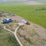 Aerial view of a rural property with a large white house, a red barn, a circular driveway, scattered trees, and expansive green fields stretching into the distance.