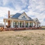 A white farmhouse with a wraparound porch, brick foundation, metal roof, and rocking chairs sits on a large grassy field under a partly cloudy sky. Leafless trees are scattered around the property.