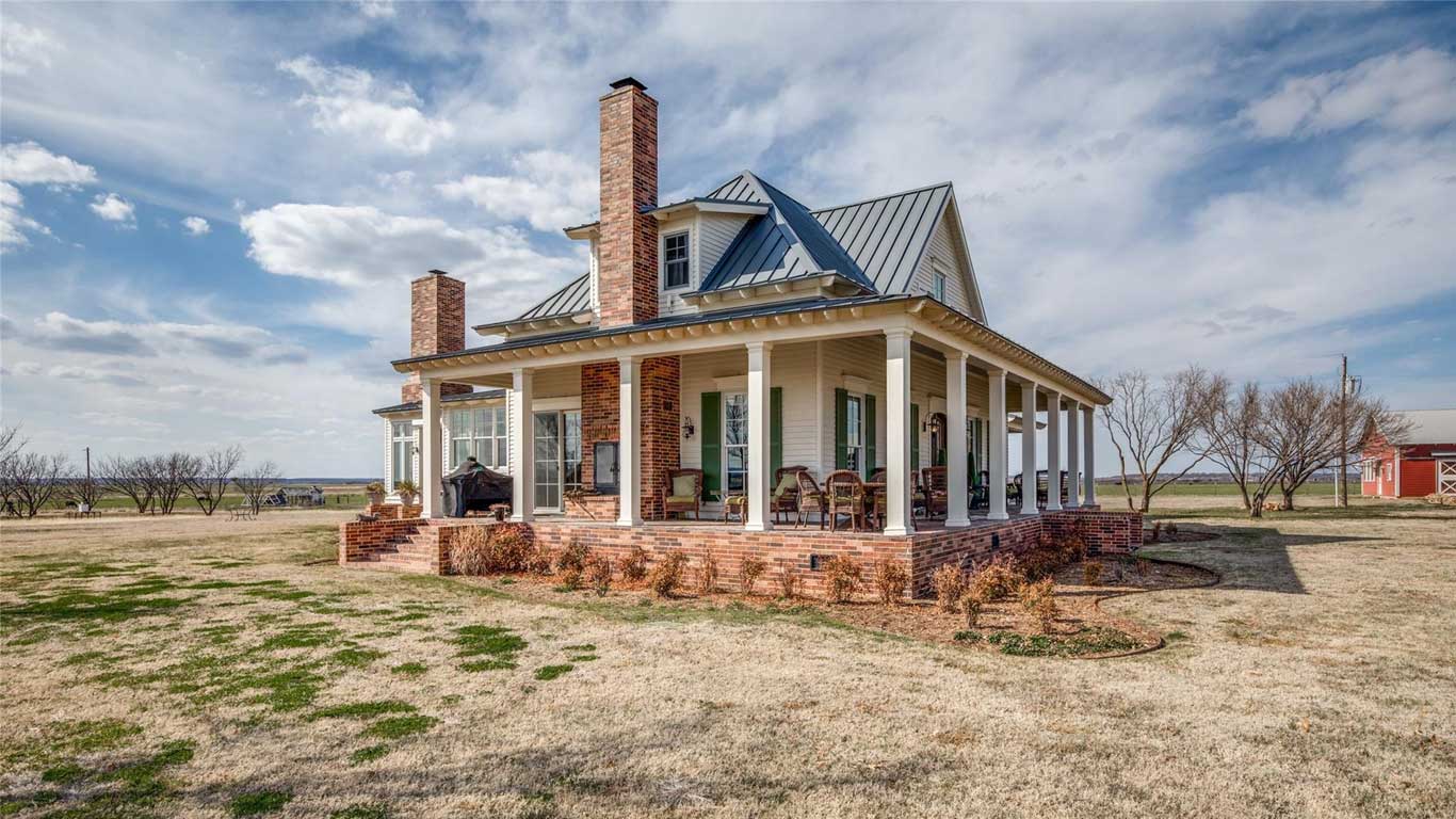 A charming farmhouse with a large wraparound porch, brick chimneys, and metal roof sits on a spacious, grassy property under a partly cloudy sky. There are rocking chairs on the porch and leafless trees nearby.