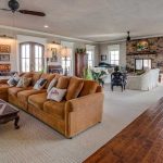 Spacious living room with large windows, brown sofa, cream armchairs, stone fireplace, wooden floors, rugs, and eclectic decor including plants, paintings, and antique-style furniture. Natural light fills the cozy space.