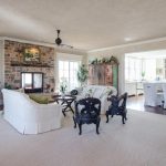 Spacious, bright living room with white sofas, dark accent chairs, a stone fireplace, grand piano, and open layout leading to a white kitchen and dining area with natural light from large windows.
