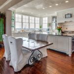 Bright, spacious kitchen and dining area with white cabinetry, marble countertops, a rustic wooden table with white slipcovered chairs, hardwood floors, large windows, and green accents.