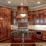 A luxurious kitchen with dark wood cabinets, a large central island with a sink, granite countertops, stainless steel appliances, and a decorative range hood beneath a hanging light fixture.