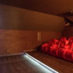 A private home theater room with red recliner chairs arranged in rows, facing a large screen on the left wall. The lighting is dim, and the ceiling is sloped. The floor is carpeted and a subtle aisle is lit along the front.
