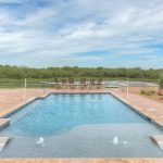 A rectangular outdoor swimming pool with two in-water loungers, surrounded by a stone patio. There's a covered patio on the left and a dining table with chairs in the background, overlooking a grassy field and trees.