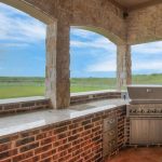 Outdoor kitchen with stone and brick countertops, built-in stainless steel grill, sink, and storage cabinets, under a covered patio overlooking a large, grassy field and distant trees.
