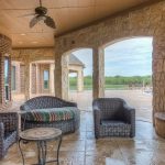 Covered patio with wicker furniture, a round table, a stone fireplace, and a mounted TV. Arched stone openings reveal a green landscape and blue sky in the background. Ceiling fan overhead.