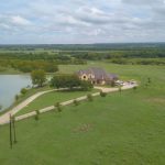 Aerial view of a large house with a driveway, surrounded by green fields, trees, and a pond under a partly cloudy sky.