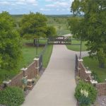A concrete driveway lined with brick pillars and landscaping leads through a gate reading "Serenity Ranch," surrounded by green grass, large trees, and open countryside under a partly cloudy sky.