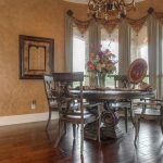Elegant dining room with round wood table, four chairs, decorative centerpiece, framed wall art, chandelier, hardwood floor, and large windows with drapes allowing natural light into the space.