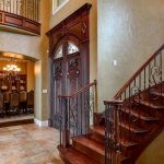 A grand foyer with a curved wooden staircase, ornate iron railings, double wooden doors, and stone tile flooring. An elegant dining room with a chandelier and large table is visible through an arched doorway.