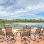 Six patio chairs surround a stone fire pit on a terrace overlooking a peaceful pond, with green grass, trees, and a partly cloudy sky in the background.