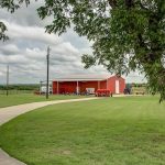 A red barn sits at the end of a curved concrete driveway on a large, well-maintained grassy property with scattered trees under a cloudy sky.