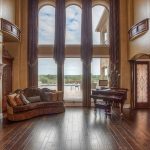 Spacious living room with tall arched windows, wooden floors, stone fireplace, elegant sofa, and grand piano, opening to a patio with scenic outdoor views. High ceilings and decorative columns add a luxurious touch.
