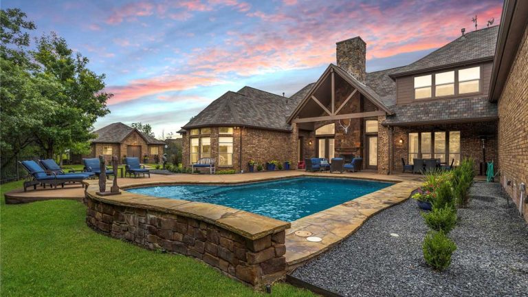 A backyard with a stone-edged swimming pool, lounge chairs, and a garden, set beside a large brick house under a colorful sunset sky.