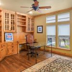 A home office with built-in wooden shelves, glass cabinet doors, a glass desk, ornate wooden chair, large rug, ceiling fan, and two tall windows with white blinds letting in natural light.